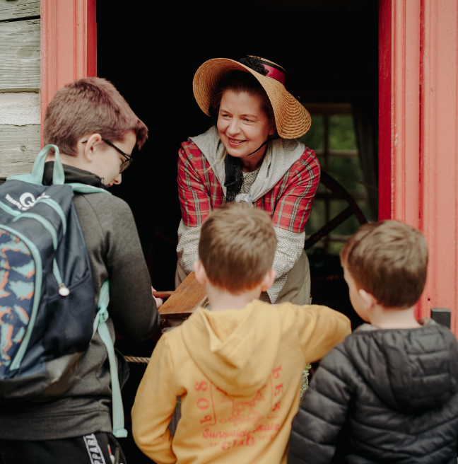 A woman, smiling, interacting with children - appears to be giving something out to him from a doorway - she looks like she's dressed like she lived on the American Frontier back in the 18th century. It's three boys, on with a backpack on and wearing glasses, the boy in the middle wears a yellow jumper and the boy on the right is in a black coat.