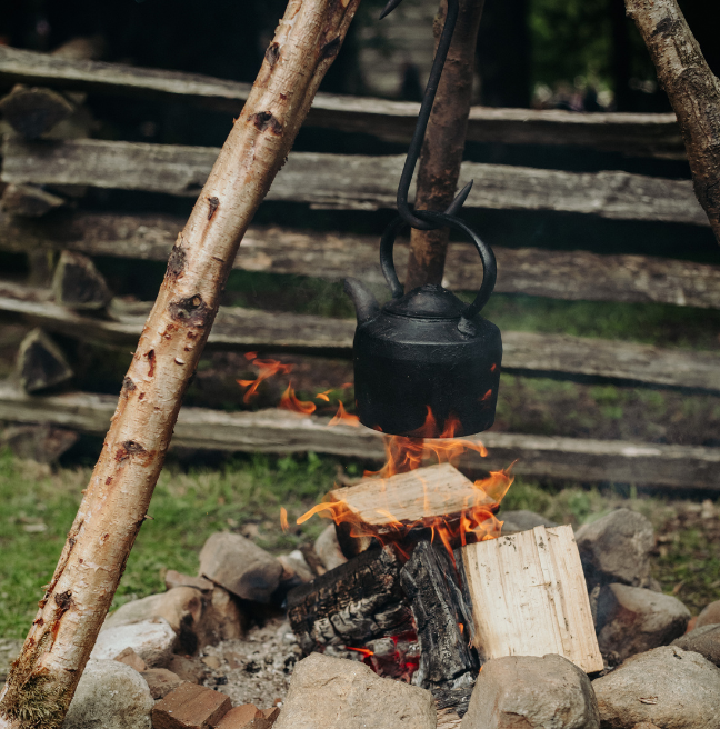 A wooden log tripod fire, with a black teapot hanging over the lit fire. 