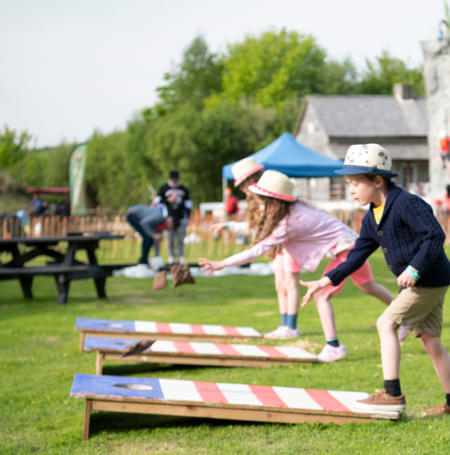 Kids playing in a grassy area, throwing bean bags onto a red, white and blue painted wooden board, aiming for the hole in the board. All three kids are wearing shorts and sun hats.