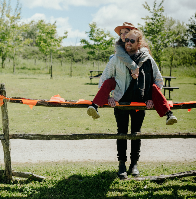 A guy and a girl at a music festival - he's standing at a fence with orange bunting on it, wearing sunglasses, and the girl is on his back, like a piggy back, and she's wearing a cowboy hat.