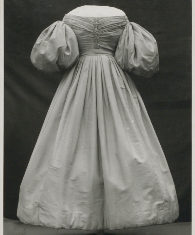 A black and white photograph of a long gown, with a low straight neckline and big puffy half sleeves.