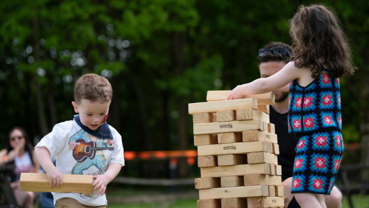 Two children with their dad playing giant Jenga outside. You can see orange bunting on a fence in the background.