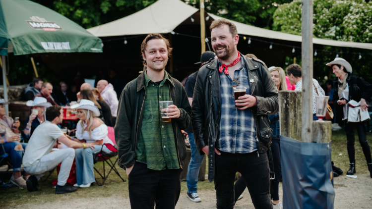Two guys wearing jackets and shirts, standing in front of an outdoor bar, holding pints. More people are seen in the background, seated in the outdoor bar, under outdoor umbrellas.