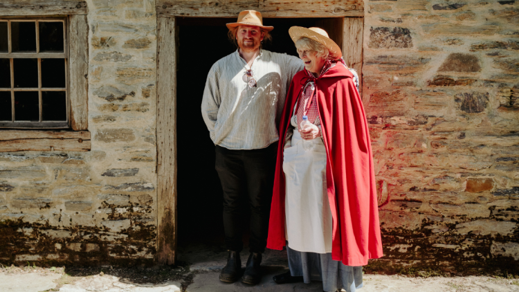 A visitor wearing a cowboy hat interacts (laughing) with a costumed visitor guide (wearing a red cloak). He has his arm around her shoulders.