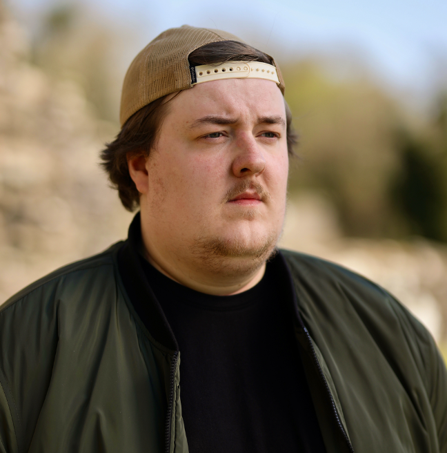Gareth Hamilton, musician, wearing a baseball cap backwards, a black tshirt and green jacket