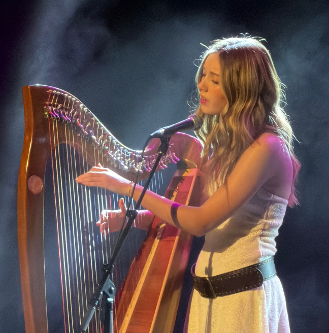 Niamh Noade, a young woman with blonde hair, playing a harp and singing into a microphone