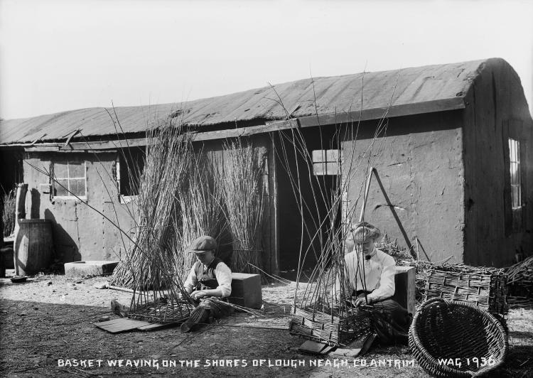 A black and white photograph of two men sitting weaving baskets outside of a barn-like workshop.