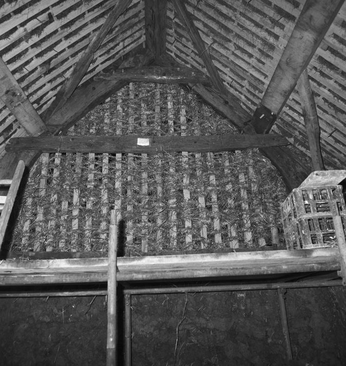 A black and white photograph showing a traditional wall construction. The upper frame is being filled with hay.