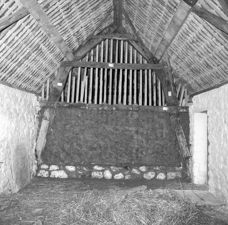 A black and white photograph showing a traditional wall construction. The upper frame is being filled with hay.