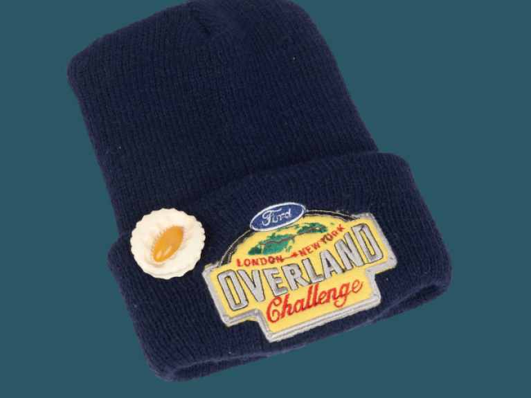 A blue beanie hat with a patch that reads 'Ford London - New York Overland Challenge'. There is a brooch shaped like an oyster attached.