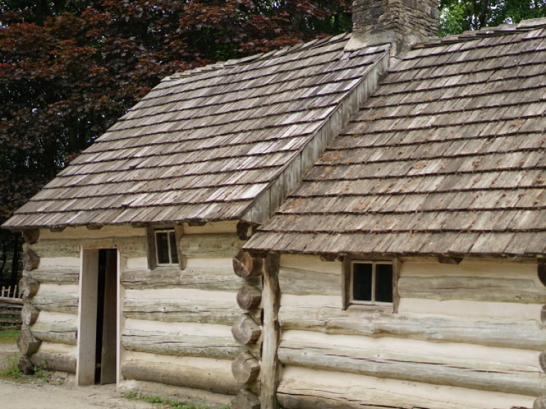 A log cabin, made of two rooms. Behind the cabin are large trees. To the left, the leaves are brownish red; to the right, they are green.