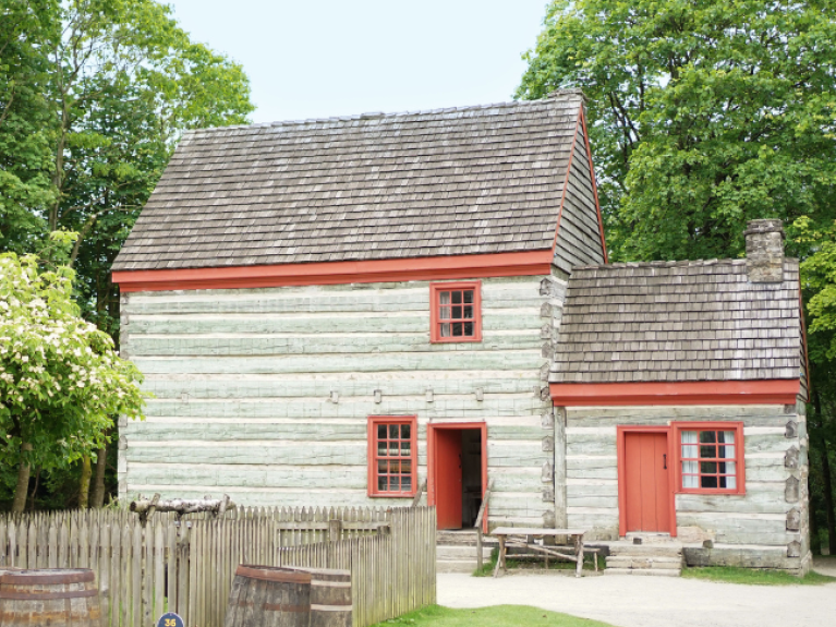 A log farmhouse with two storeys sits amidst some trees. The logs are varyingly blue and grey, while the trim is red.