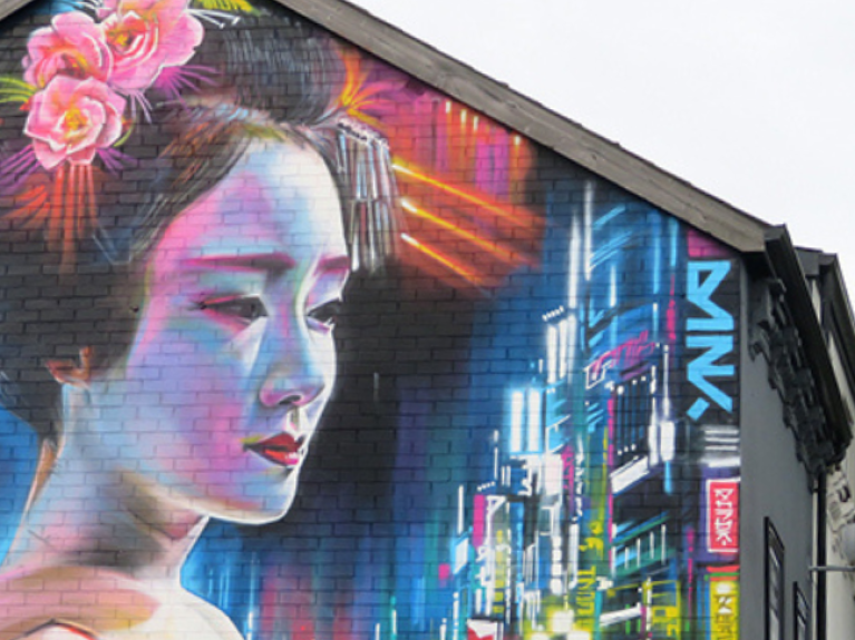 An image of a mural on the end of a building. The mural is of a Japanese woman, wearing her hair up with flowers. In the background is a vibrant, neon city-scape. The lights behind her head look almost like a halo. 