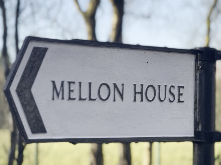 A sign outdoors shaped like an arrow, reading 'Mellon House'. 