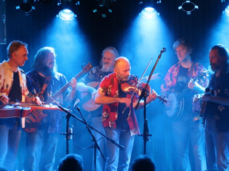 a six piece male band on stage under blue light playing instruments, band is called rawhide