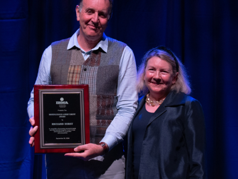 A man dressed in a shirt and waistcoat holding an award plaque beside a smaller in height woman, with shoulder length fair hair wearing a black outfit. There is a blue curtain as the backdrop.