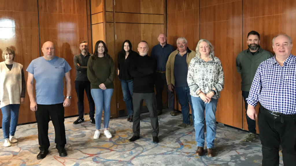 Group of 11 people standing in a carpeted room and wood panelled walls. Mixture of men and women who have formed a group called The Conflict and Legacy Interpretive Network