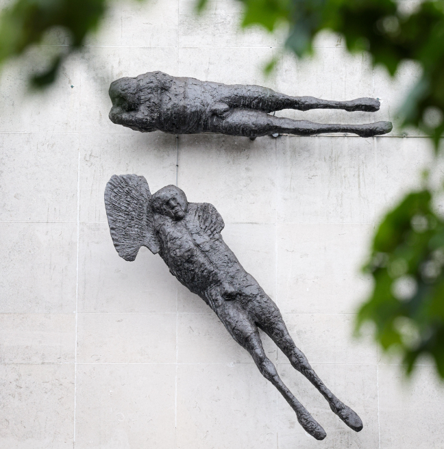 Two grey, aluminium sculptures by Elisabeth Frink attached to a light grey wall, with greenery of a tree in shot also.