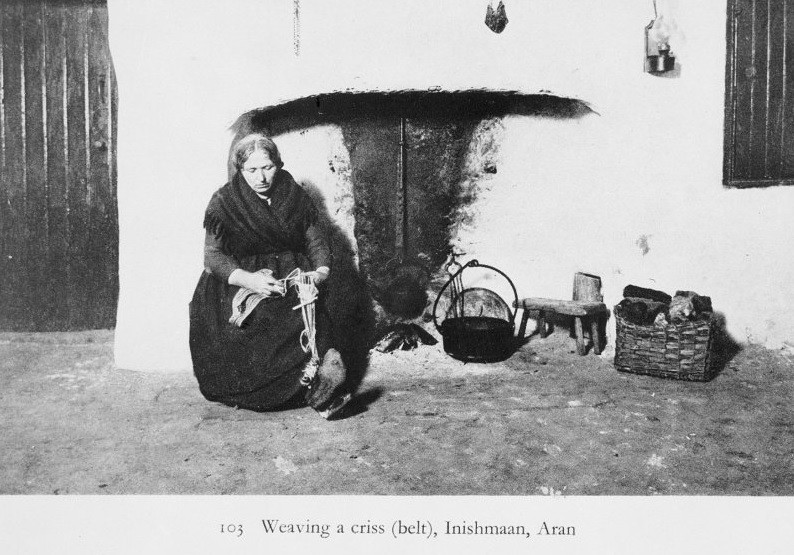 Black and white photograph of an older woman weaving by the fireplace
