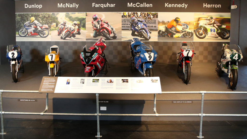 Six motorcycles displayed on a grey slanted platform with pictures of the racers that rode them on wallpaper on the wall behind each one. From left to right the wallpaper says Dunlop, McNally, Farquhar, McCallen, Kennedy and Herron.