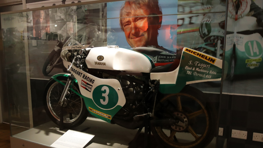 A green and white bike that once belonged to Joey Dunlop inside a glass case on display in the Ulster Transport, with a picture of Dunlop on the wallpaper graphic behind the case.