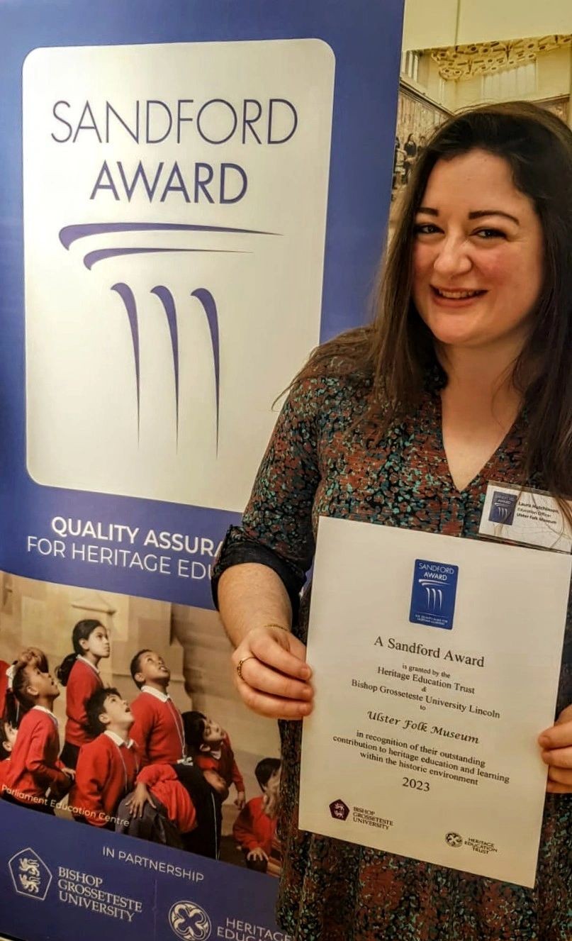 A person with long dark hair, smiling, holding a certificate award beside a banner with a blue frame that says Sandford Award.