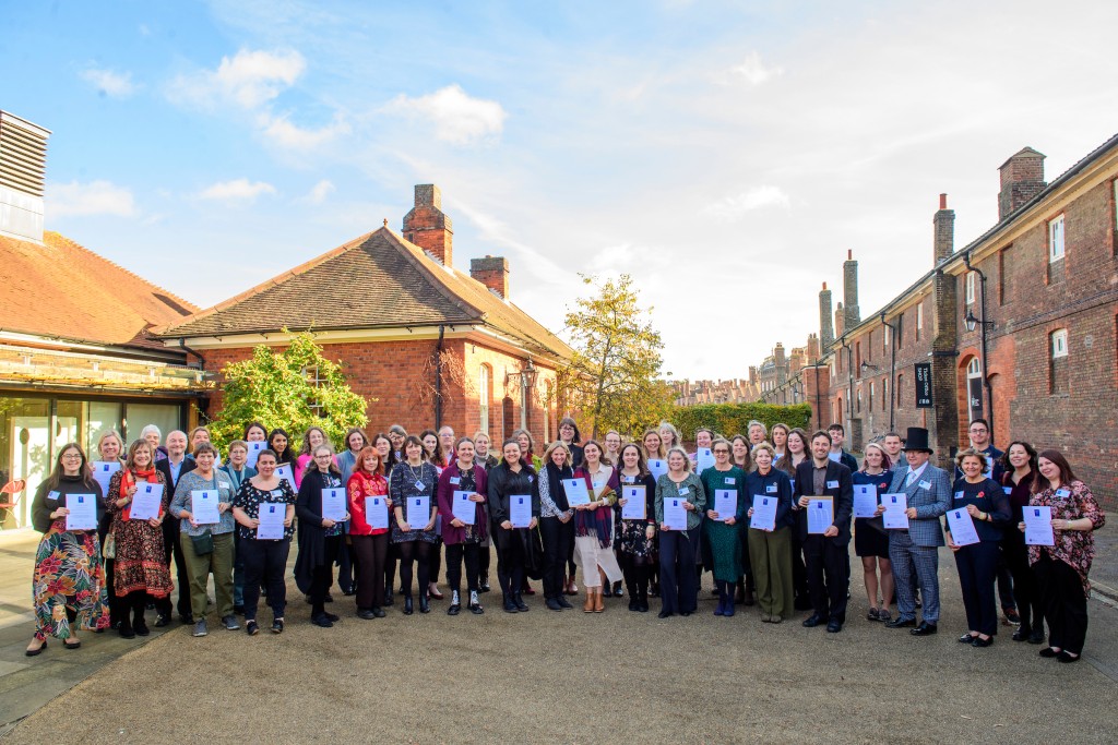 A group of people standing in a line at Hampton Court Palace, holding certificate awards. The sky is blue and the sun lights up the buildings in the background.