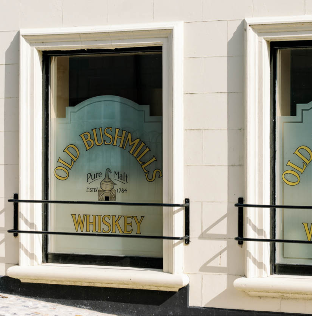A rectangular window with black metal bars across the bottom. The windows are frosted, with 'Old Bushmills Whiskey' and the Bushmills logo hand-painted into the frosting.