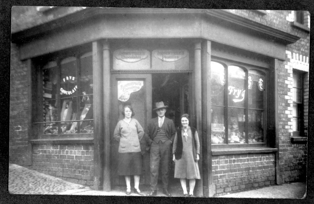 Two women and a man stand outside of a corner shop in an old black-and-white photograph. They are dressed in clothing from the 1920s. The shop behind them advertises chocolate on its windows.