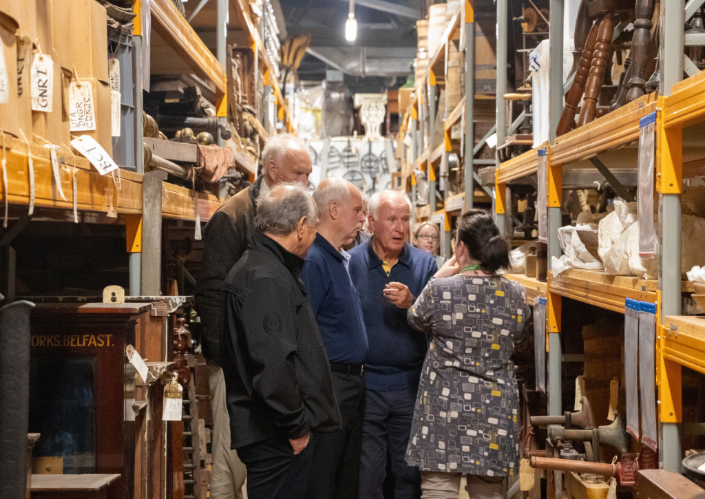 A group of people walk down the aisle of a collection store. They are gathered around a curator, who is speaking