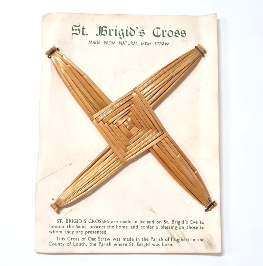 A St Brigid's Cross mounted on cardboard. The cardboard notes details about the cross. The cross is golden, with a diamond shaped centred and four thick arms eminating from the sides.
