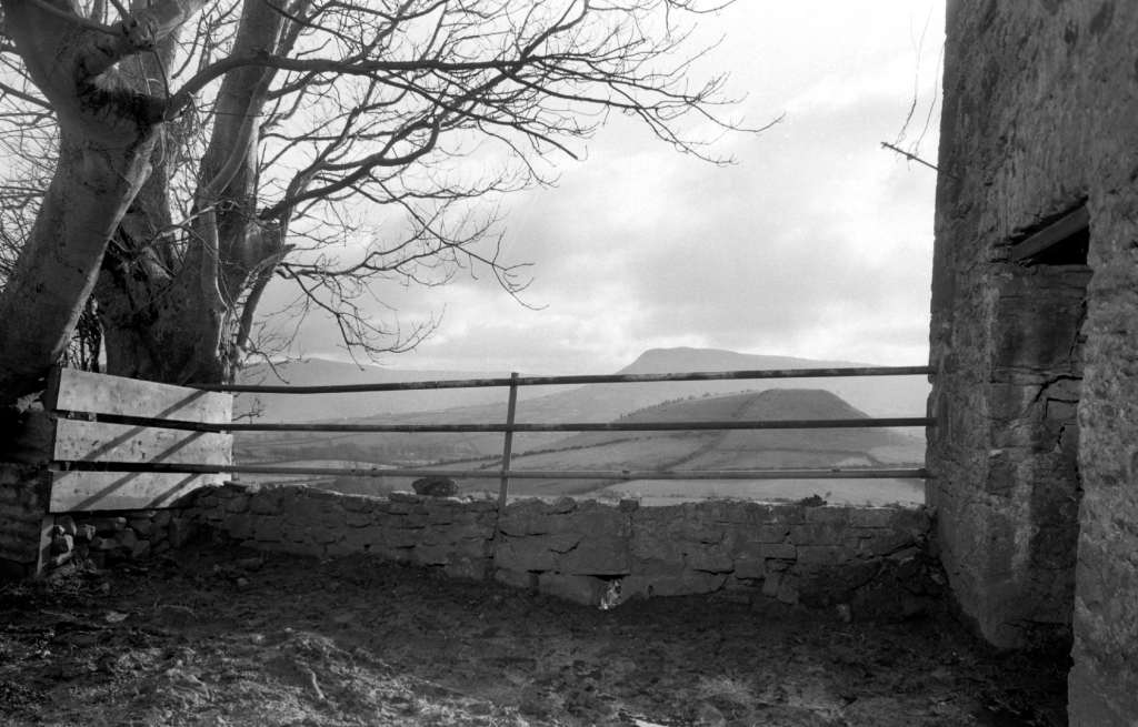 A black and white photograph taken from behind a metal fence. In the backgrounf, sccenic mountains rise into a cloudy sky.