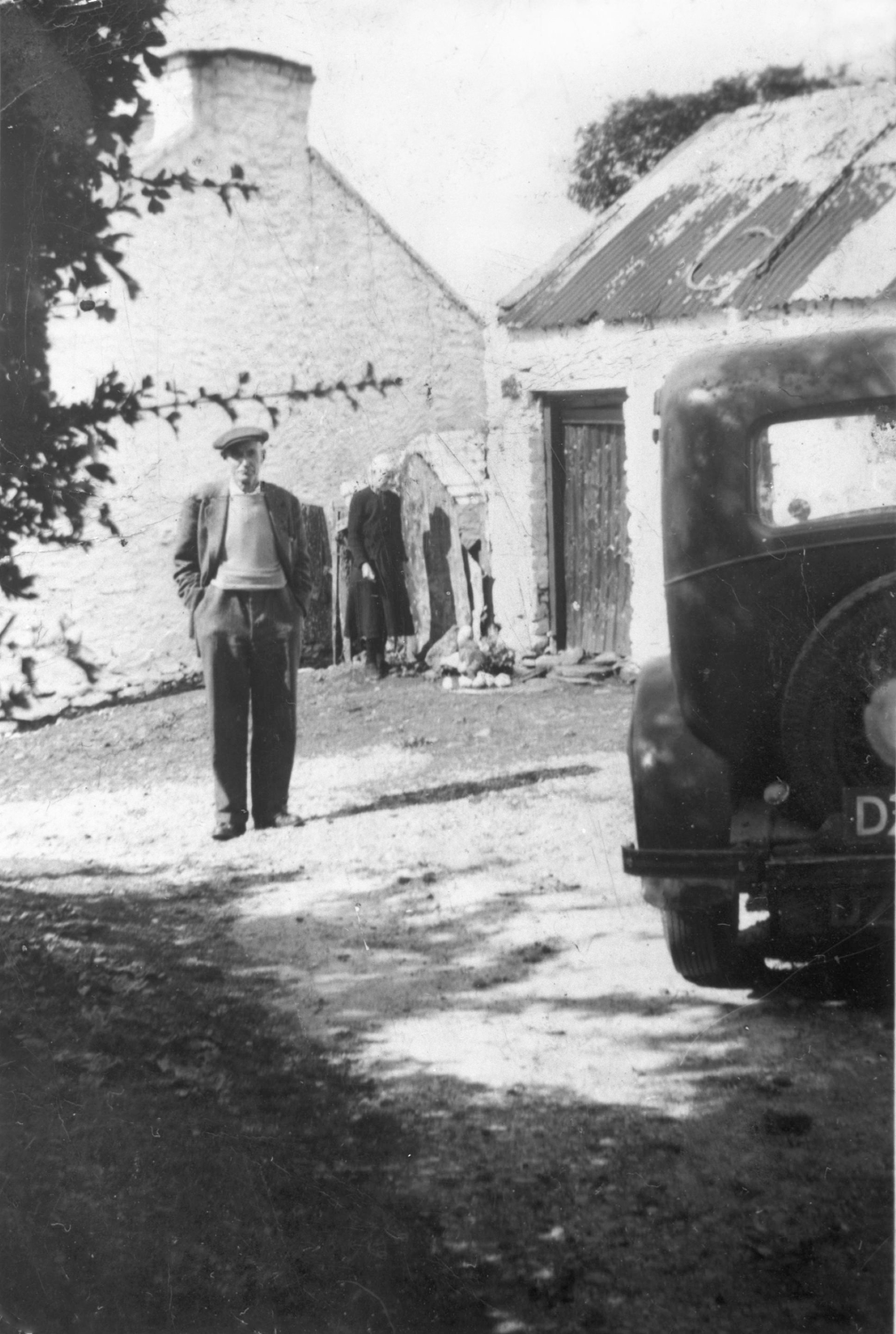 A black and white image. In the foreground is the nose of an old car. In the background, parts of two white farmhouses are seen. A man dressed in the clothes of the 1920s/30s is seen standing in front of the buildings. In the rear, a woman stands next to the buildings. 