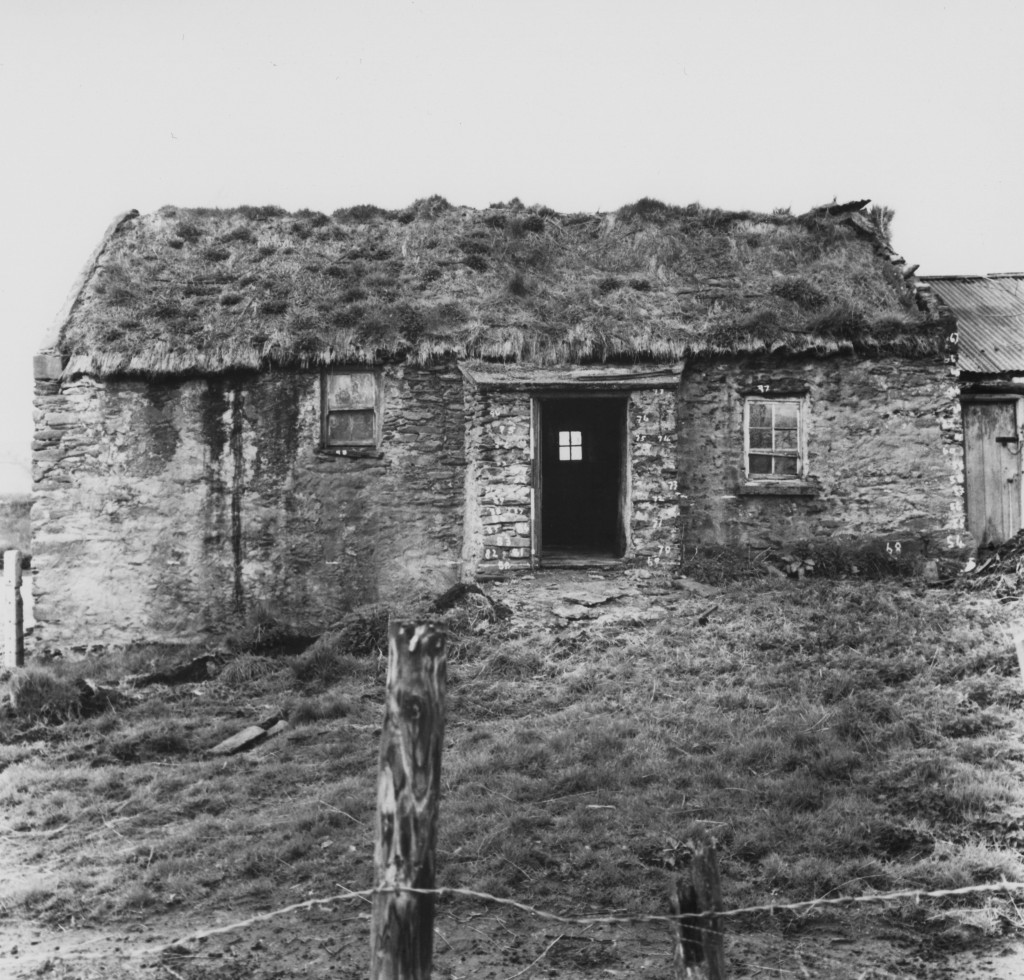 A black and white photo of a dilapitated cottage. The thatch is thinning and messy.