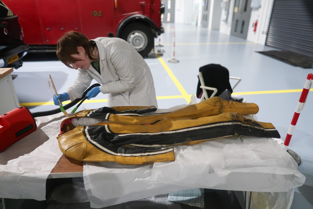 A woman stands over a yellow and black racing jumpsuit. She is assessing the top part for conservation measures. She is holding a small vacuum.