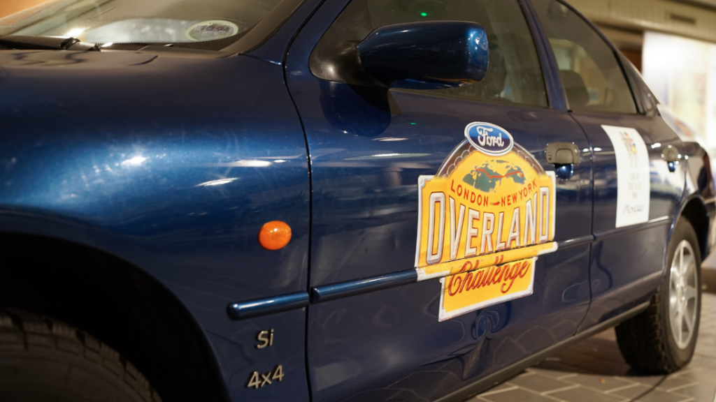 The side of a blue Ford car with a large yellow decal reading ''Ford - London-New York - Overland Challenge'.