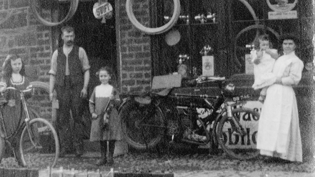 A black and white old photograph of a family, wearing somewhat Victorian/Edwardian clothes, standing in front of a bicycle shop.