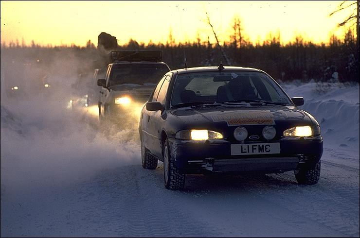 In the setting sun of a snowy outdoors, a car leads a back of cars and trucks. 