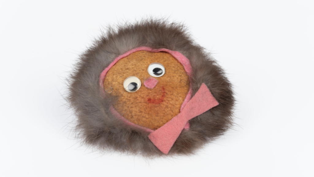 A child's craft. The object is a badge of a child's face, wrapped up warm in a fur hood with a pink bow beneath the chin. The face is childlike, with googly eyes for eyes and a simple red thread making a smile.