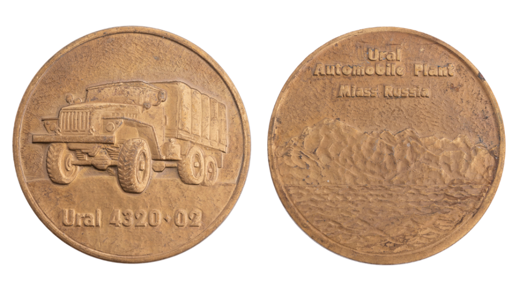 The front and back of a round copper coin. On the front is an embossment of a big truck, and on the back are some mountains and the words 'Ural Automobile Plant, Miass, Russia'.