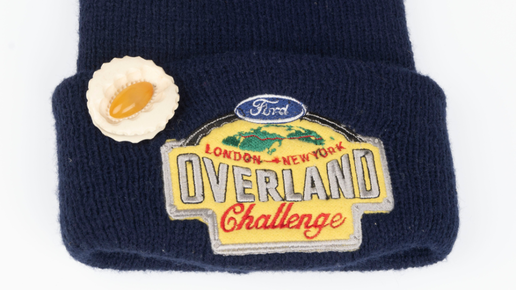A close up of a navy blue beanie. The beanie has a badge on it that looks like a flower or a seashell. The logo is a bright yellow, reading 'Ford London-New York Overland Challenge'.