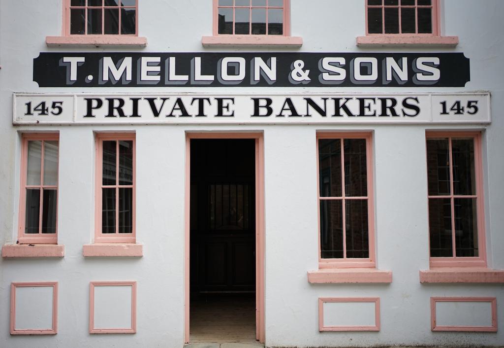 The exterior of a bank, with a large sign above the door reading 'T. Mellon & Sons, Private Bankers'