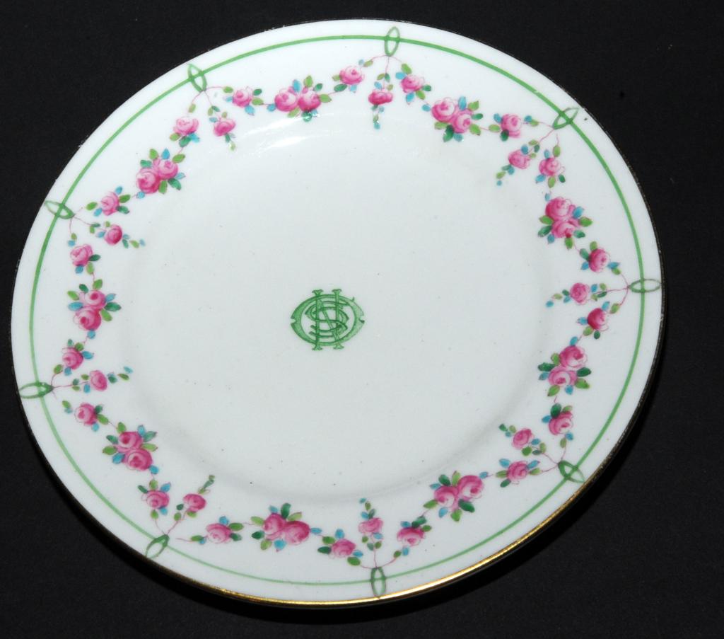 A white side plate with a rose pattern on the edge. In the centre, green letters overlap to form a stamp.