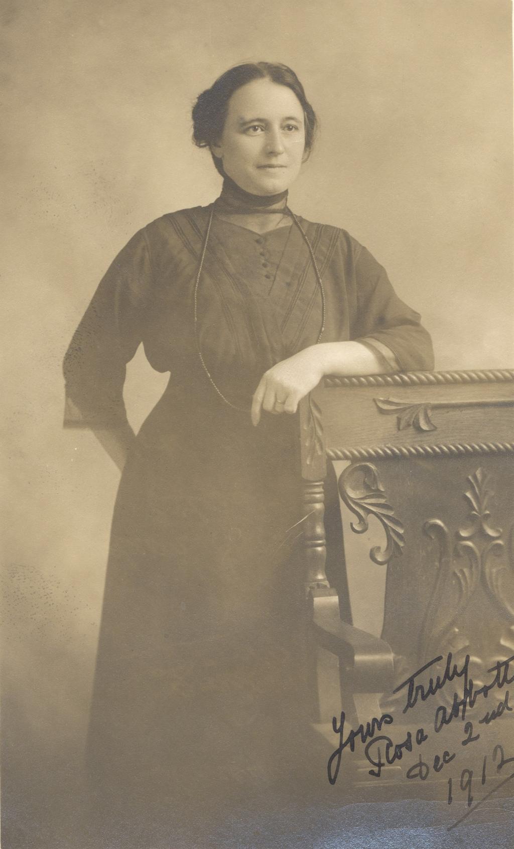 A black and white photograph of a woman standing resting on a chair. 