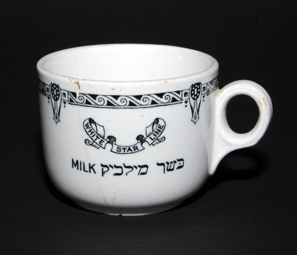 A white teacup with a black edge. The logo for the White Star Line is across the side of the cup. Beneath the logo, the word 'milk' is written in English and Hebrew.