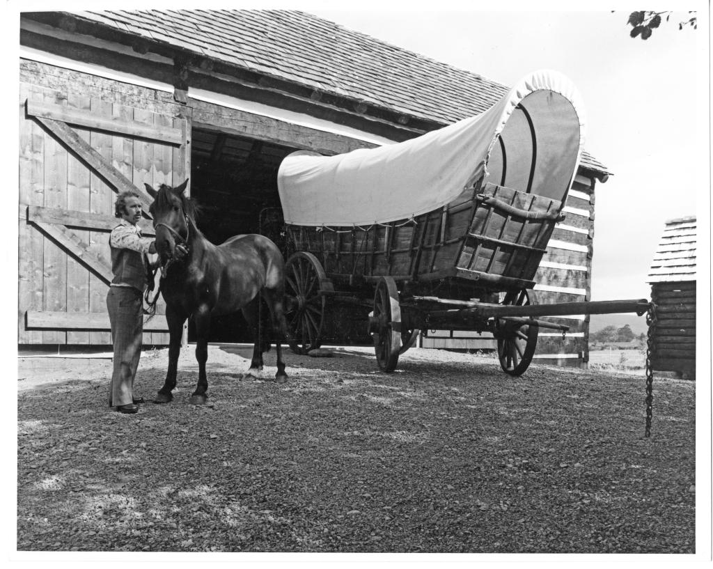 A black and white photograph of a Conestoga wago outside of a barn. A horse is standing beside it.
