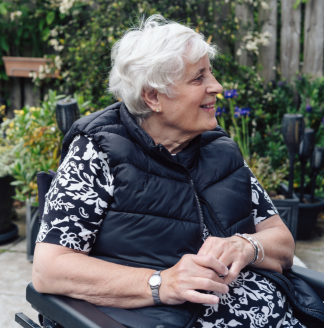 A person with white hair, sitting in a wheelchair in her garden, wearing a flowery dress and black gilet. hair. Her fingers are loosely intertwined on her lap and she has a watch on her right wrist and a bracelet on her left wrist.
