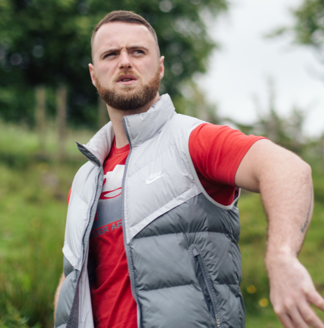 A young male with dark hair and beard standing in a grassy area of a mountain wearing grey shorts, grey gilet and a red t-shirt. His left arm is lifted away from his body, out to the side and he's looking up and into the distance.