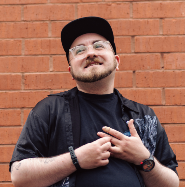 A person dressed in dark clothes, facial hair and glasses, standing against a red brick wall, smiling and looking off to the side.