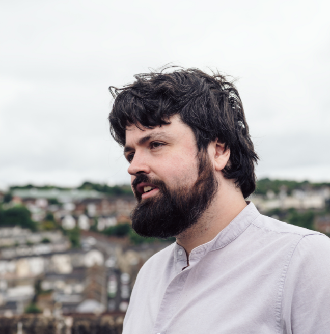 A side on, top half torso photo of a male with dark hair and facial hair, wearing a grey long-sleeved polo, with Derry city in the background